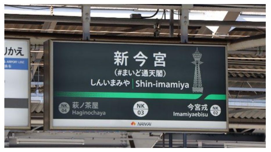 「#まいど通天閣」の副駅名が書かれた、南海電車「新今宮」の駅名表示板。通天閣のイラストも描かれている。